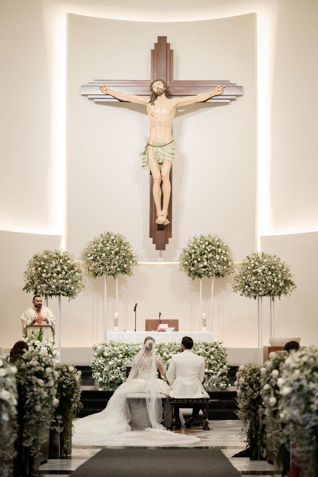 Los novios arrodillados de espaldas frente al altar, bajo el crucifijo y entre arreglos de flores blancas