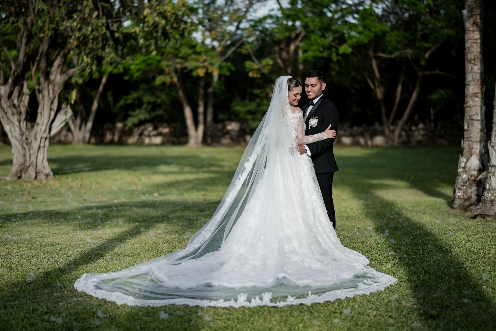 Los novios abrazados en un jardín de hacienda con sombras largas del atardecer y la cola del velo extendida