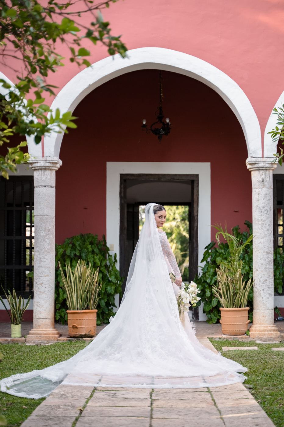 La novia con su velo largo bajo un arco blanco en el muro rojo de una hacienda